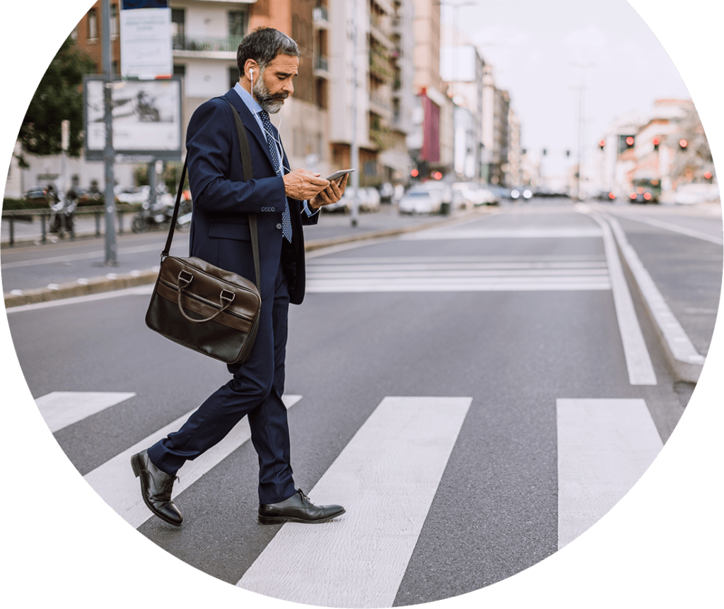 An older man in a suit is crossing a crosswalk while holding his cellphone with headphones in and carrying a messenger bag on his shoulder. Photo is inside of a circle shape.