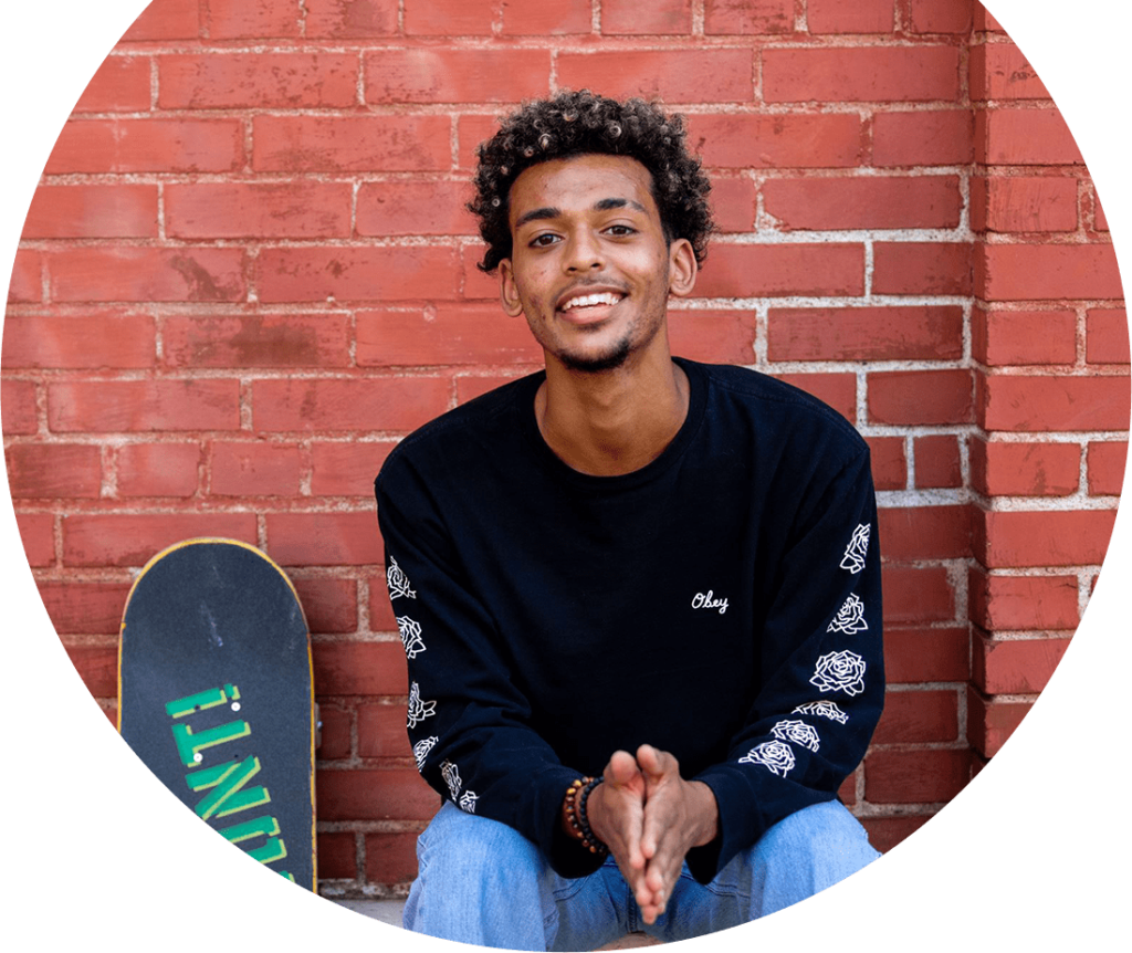 Alltru Member Andrew is smiling and sitting outside in St. Louis with his skateboard next to him in front of a brick wall. Photo is inside of a circle shape.