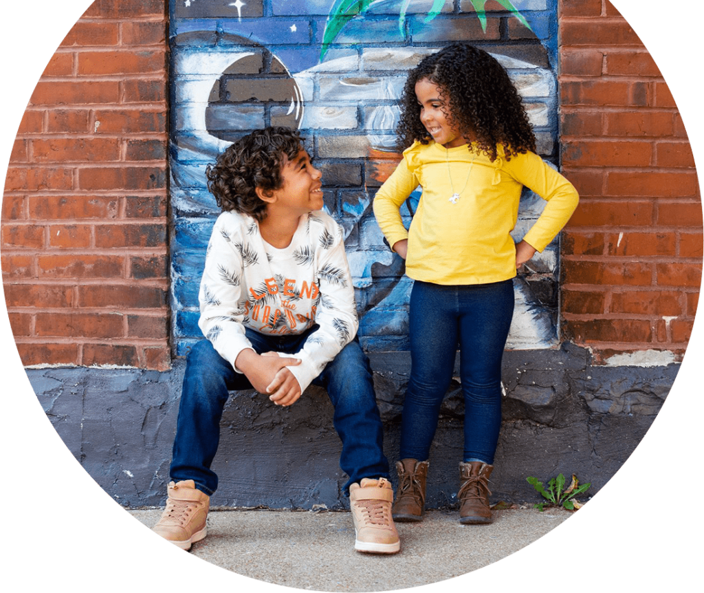 Alltru Youth Members Alijah and Emrie smile at eachother while outside in St. Louis in front of a brick wall with painting on it. Emrie has her hands on her hips while standing and smiling at Alijah while he sits on the curb. Photo is inside of a circle shape.