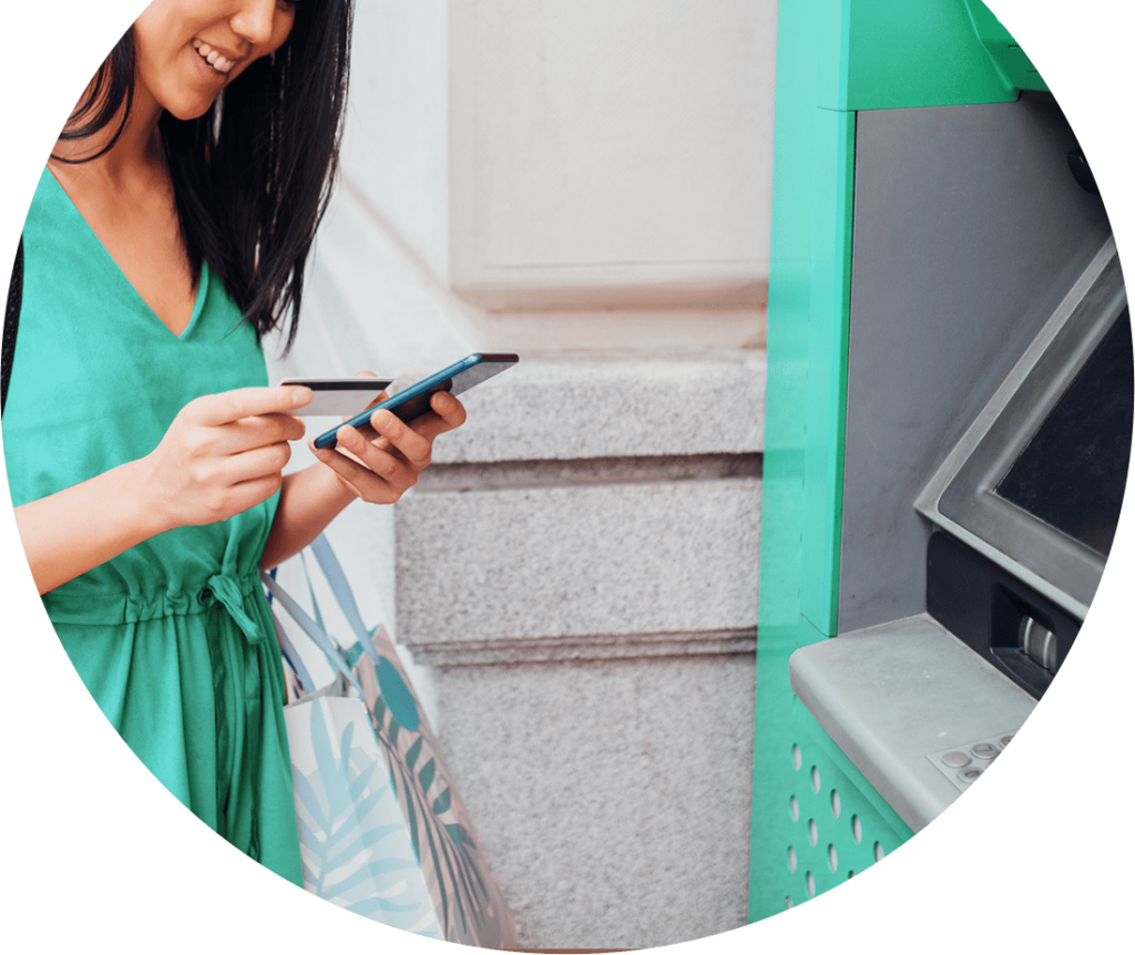 Woman standing in front of an ATM looking down at her smart phone. 