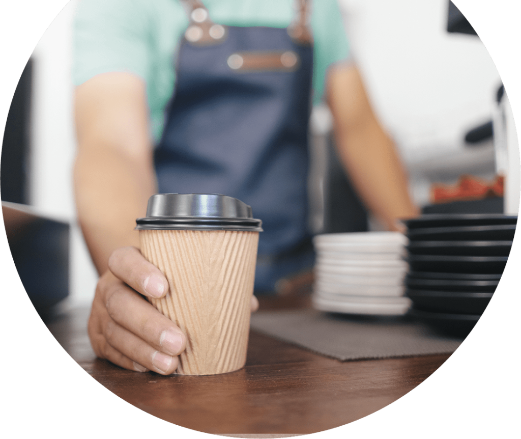 A worker at a coffee shop setting a cup of coffee on the table. Photo is inside of a circle shape.