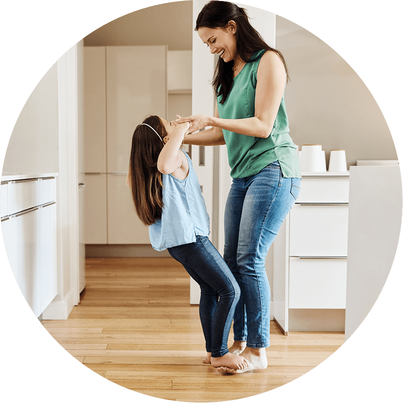 A mother and daughter are pictured dancing inside of their house and laughing. Photo is inside of a circle shape.