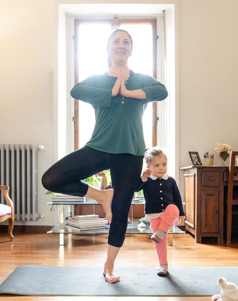 A woman and her toddler daughter do yoga together inside of a bright living room.