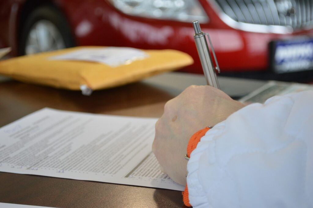 A man signing an auto loan contract with his new red car in the background