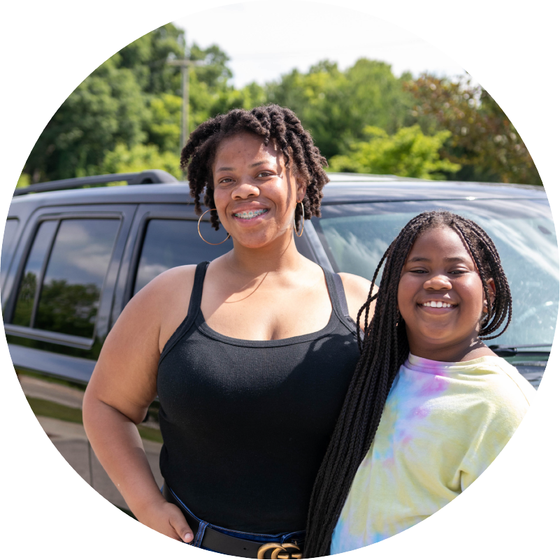 Alltru Members Tranita and her daughter Camryn standing in front on their car smiling. Photo is inside of a circle shape.