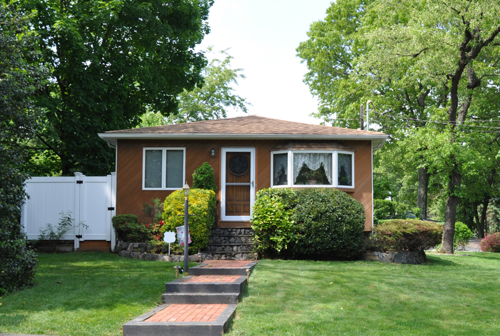 A small red-brown home with a brick sidewalk leading to the front door sitting on a green lawn with a white privacy fence to the left